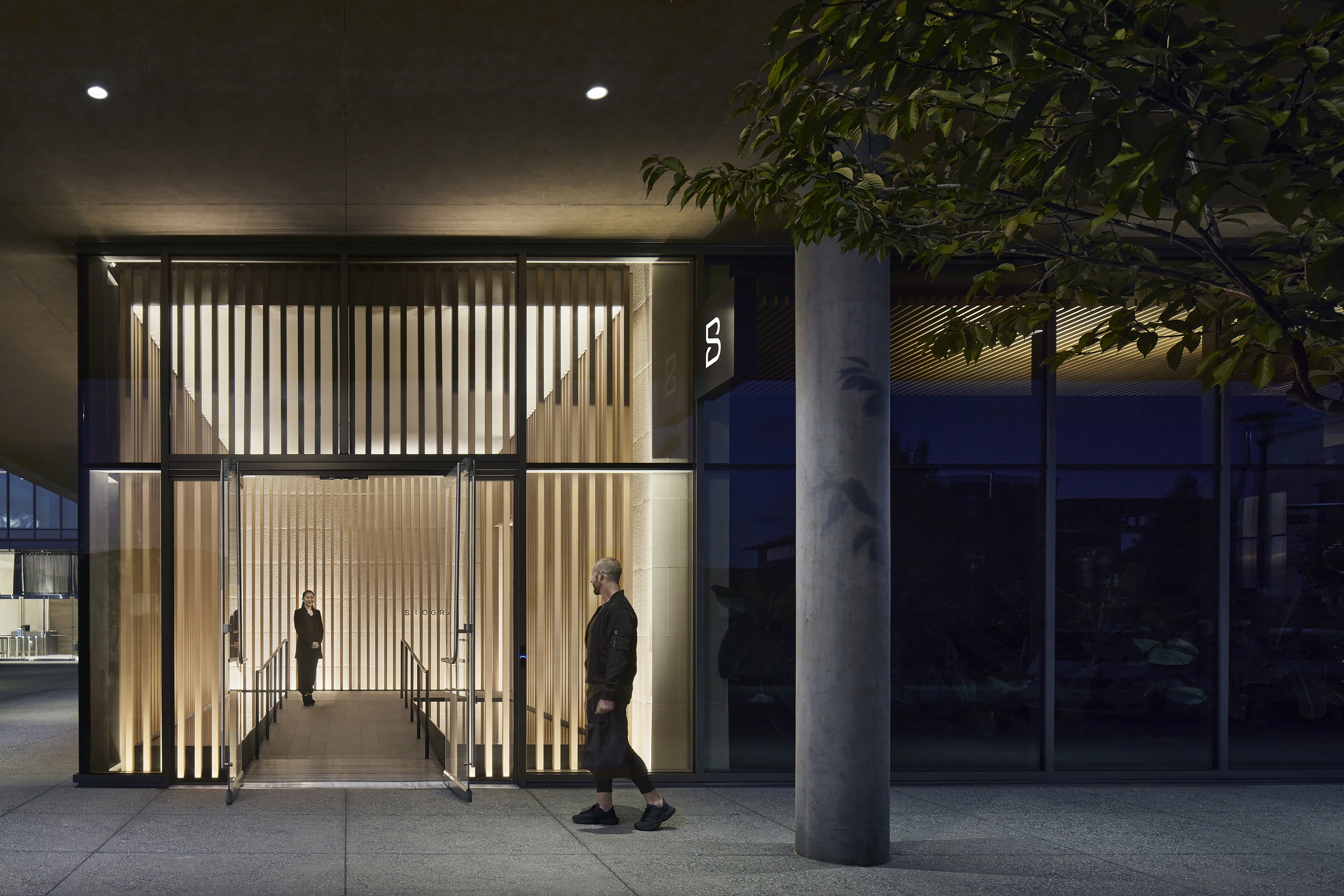 Biograph clinic entrance at night with vertical wood slats, glass facade, and a passerby.