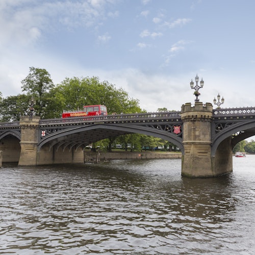 一辆红色双层巴士穿过一座华丽的桥，桥下是一条河，背景是树木和部分多云的天空。