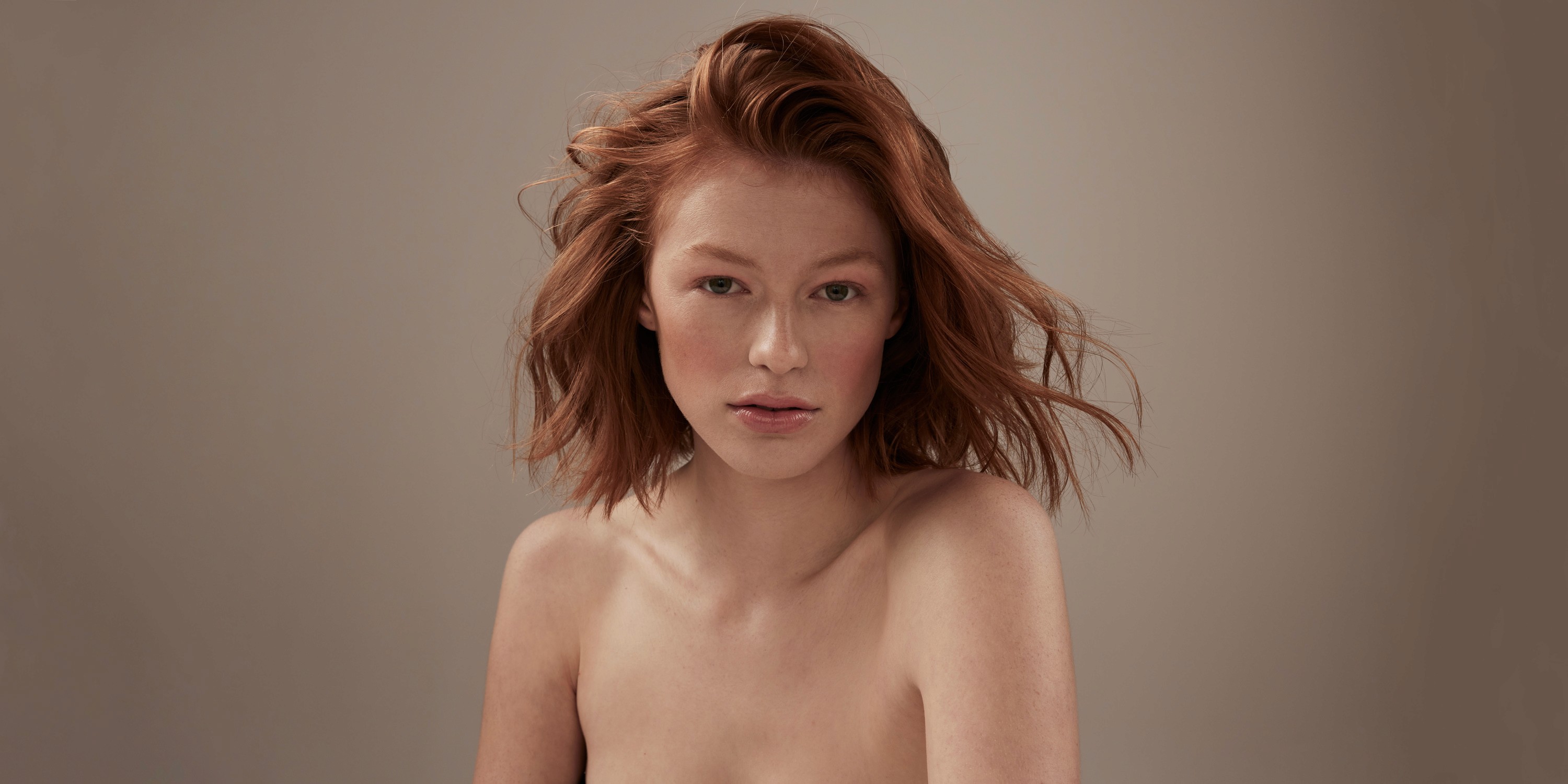 Beauty photography of a model with shoulder-length red hair and bare shoulders on a cream background.