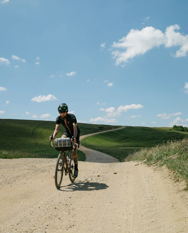Cycliste pratiquant le vélo gravel sur un large chemin de terre poussiéreux en pleine nature sous le soleil.