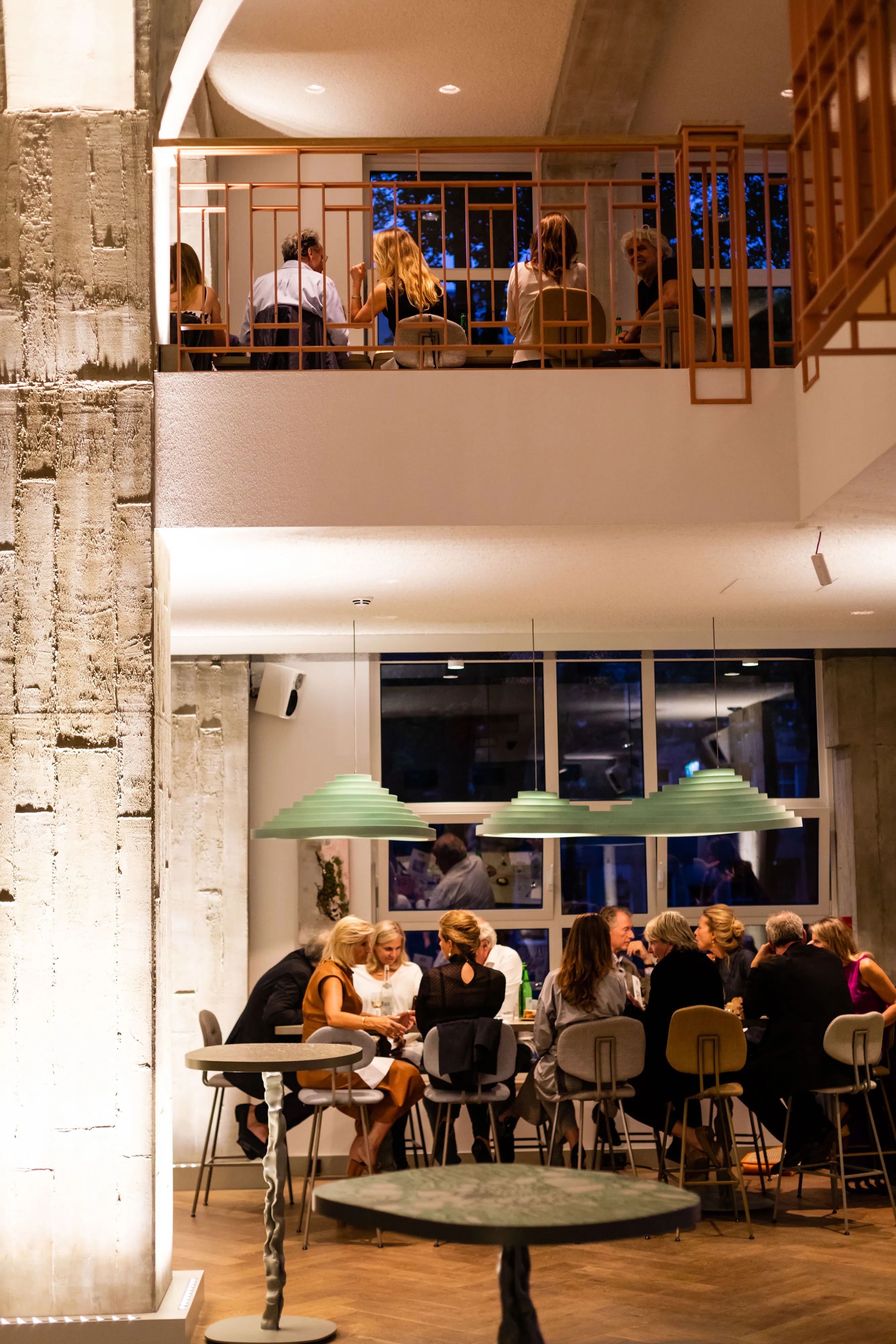 A cozy, modern cafe with customers seated at tables, some on a balcony above, illuminated by warm lighting.