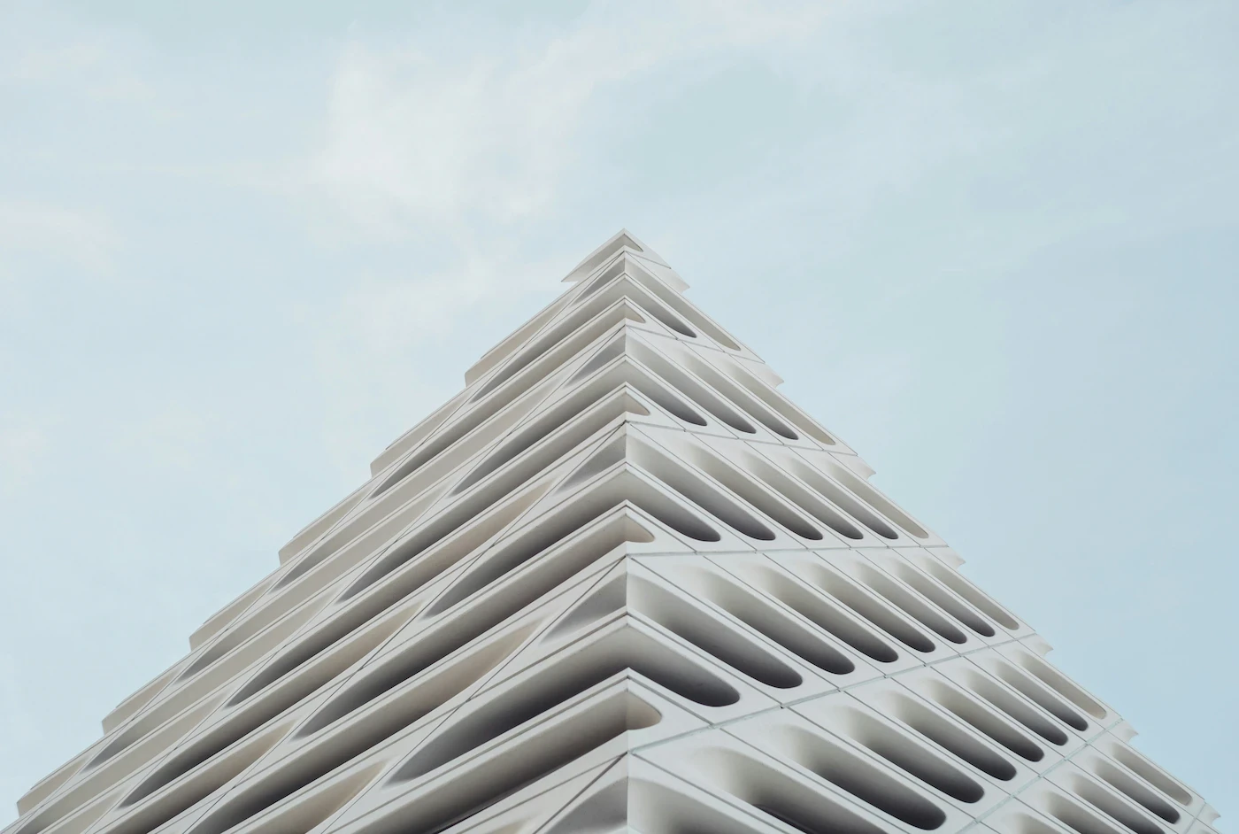 Low-angle view of a modern, multi-tiered building with a distinct pyramidal or stepped architecture, rising up toward a soft, overcast sky.