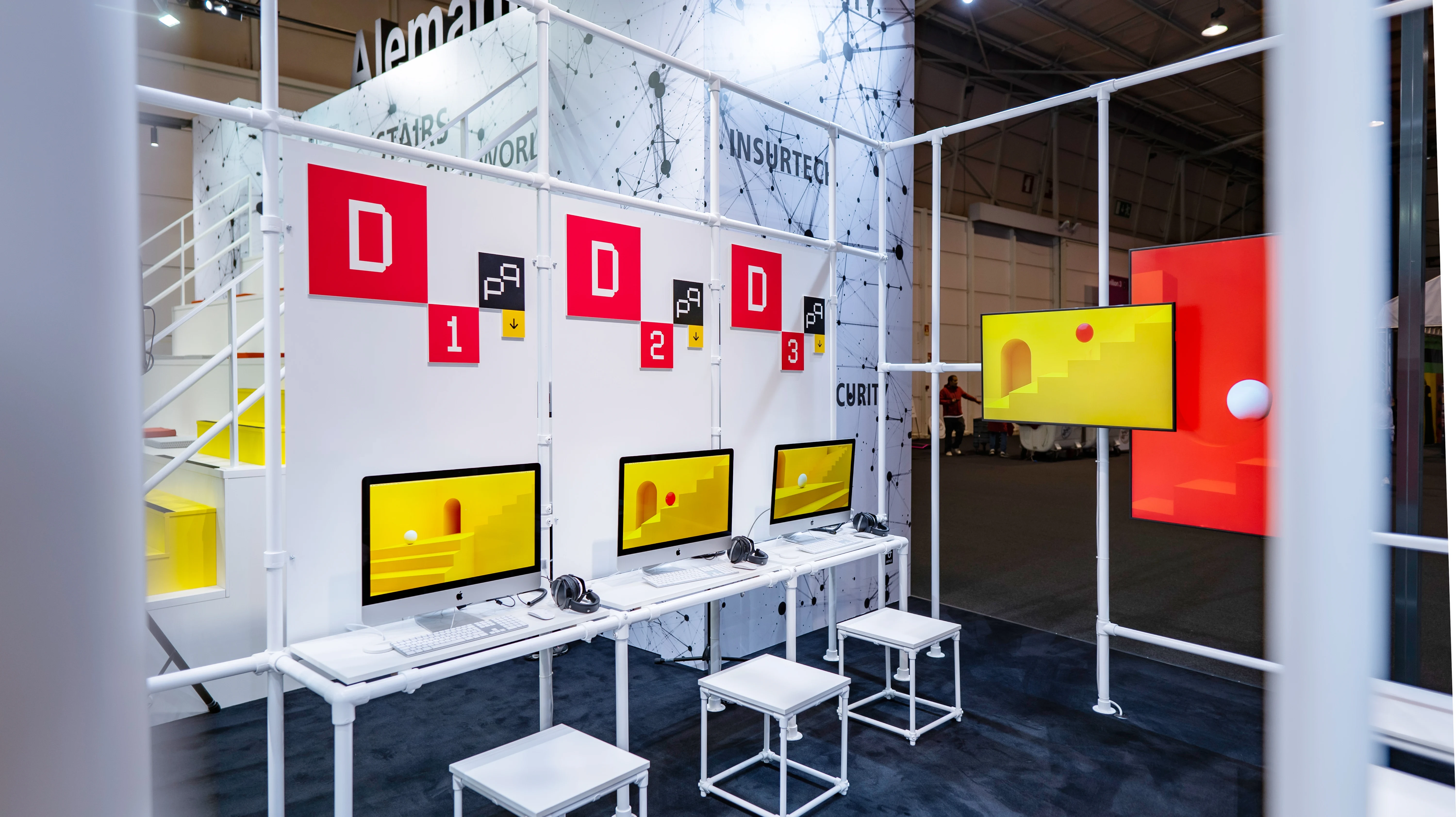 Workstations with iMacs and yellow-red screen graphics along a white frame wall at the German Pavilion at Web Summit.