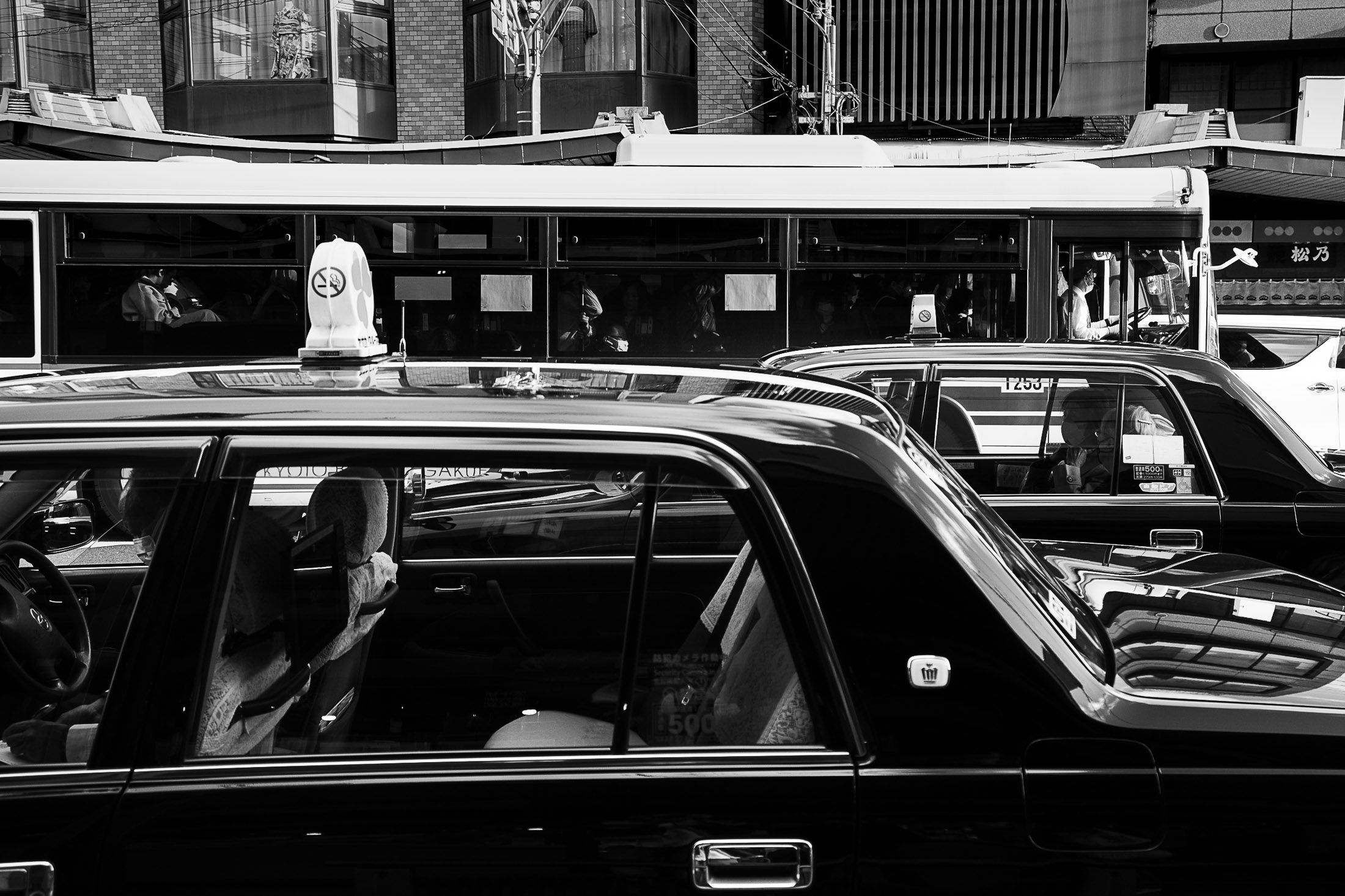 Black and white photograph of Tokyo taxis and bus with passengers seen through windows in an urban scene