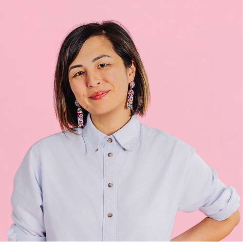 Woman with a short bob haircut wearing a light button-up shirt and colorful earrings, smiling gently against a pink background.