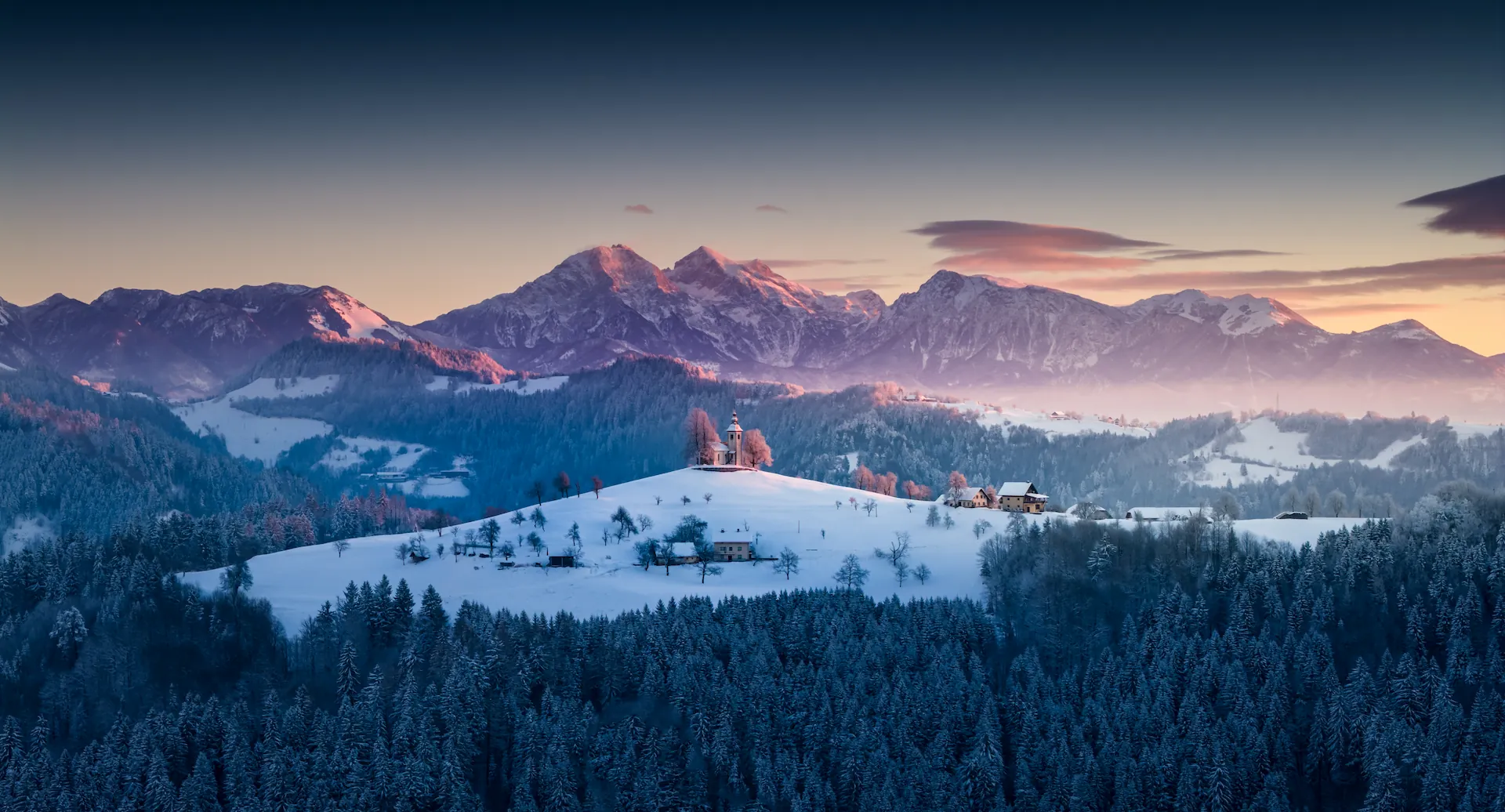 A wide winter landscape of Sveti Tomaž, Slovenia, featuring a hilltop church and scattered houses overlooking a valley of frozen trees under a soft dawn sky.
