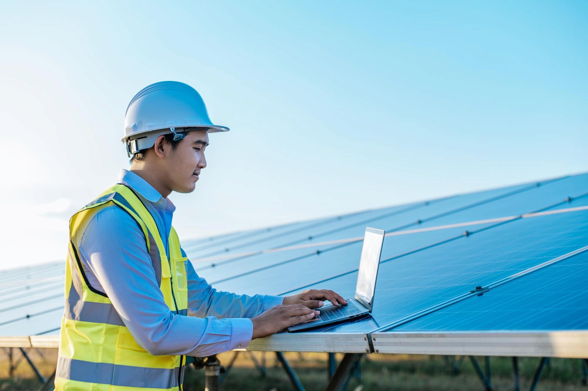 A solar energy technician in a hard hat and safety vest works on a laptop near solar panels under a clear sky.