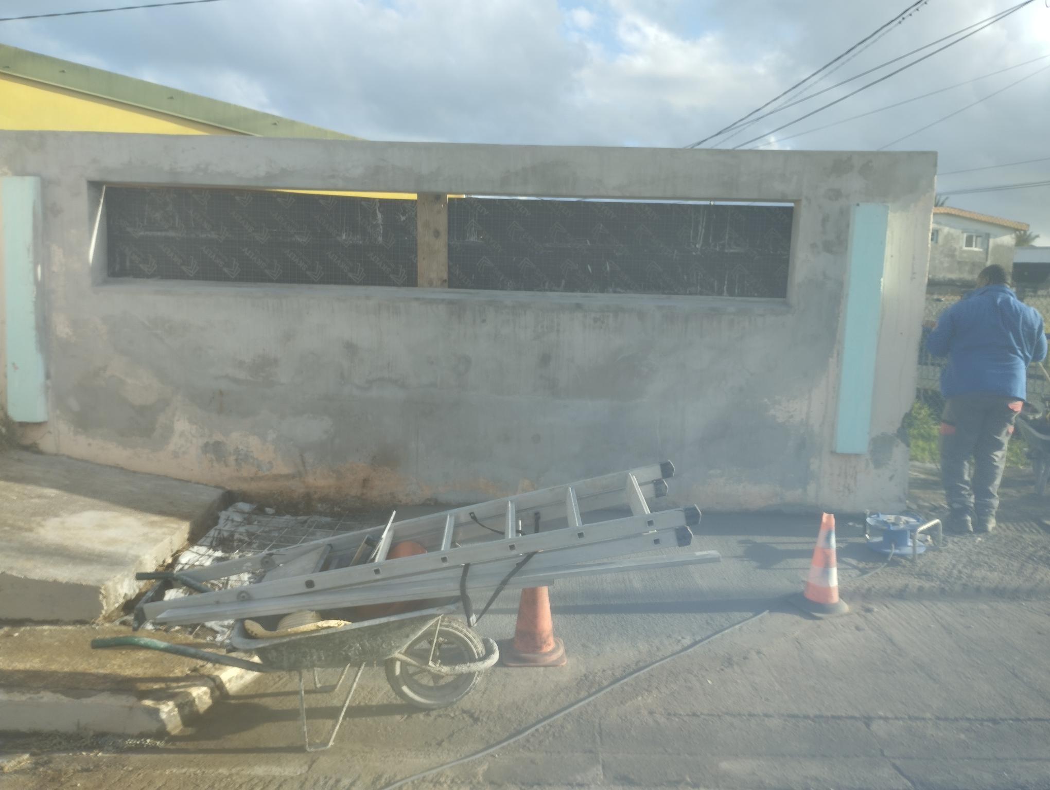 Sous un ciel nuageux, un ouvrier d'Agenov en bleu ajuste un mur en béton. Une échelle argentée repose sur une brouette. Deux cônes orange se trouvent à proximité sur le trottoir.