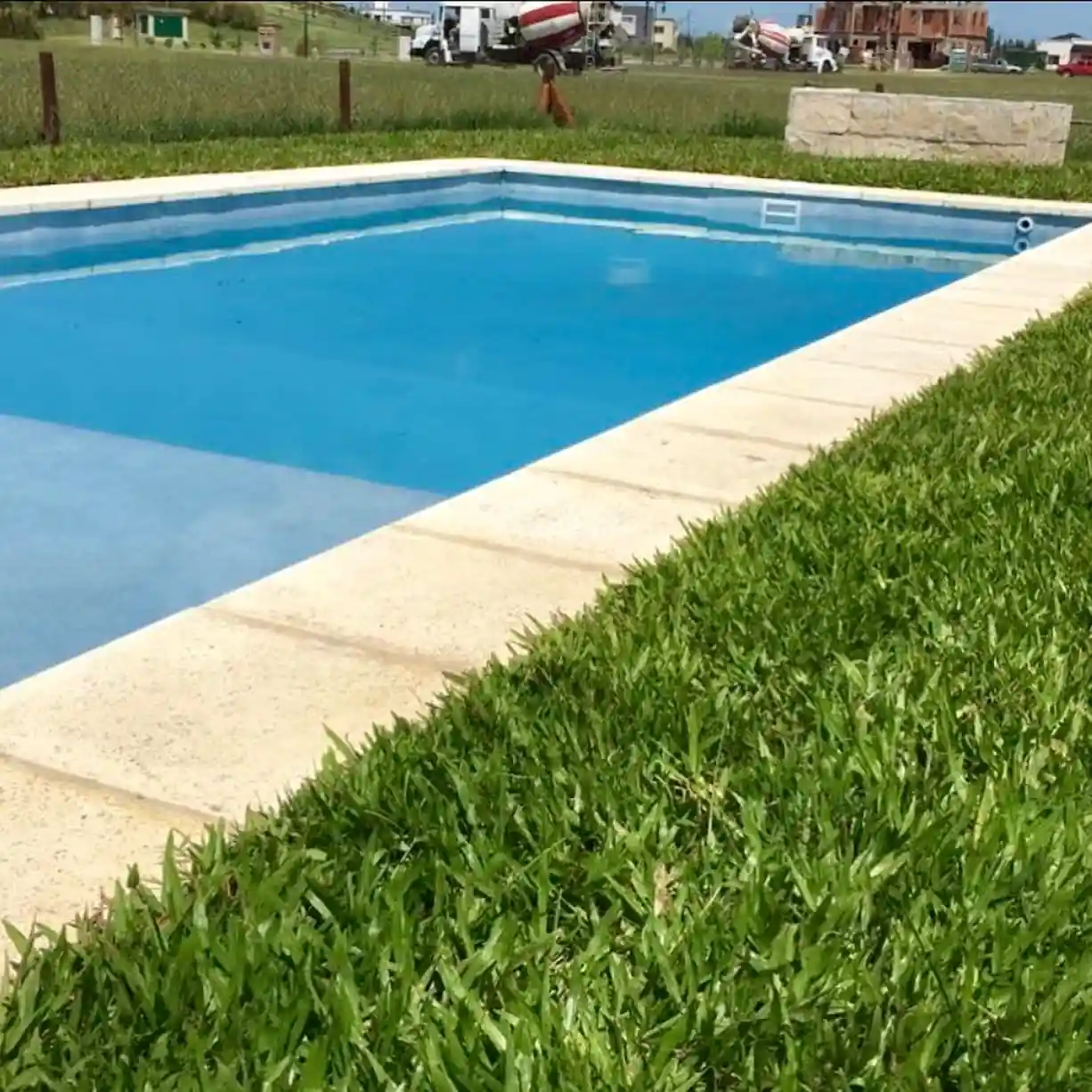 Clásica pileta terminada con agua color celeste vibrante y bordes atérmicos claros, integrada al paisaje verde de un barrio privado en desarrollo.