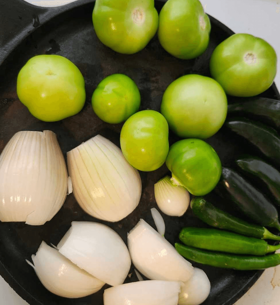 Fresh tomatillos, onions, and green chiles arranged in a cast iron skillet before roasting.