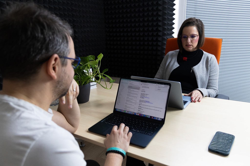 Člověk sedí u stolu a pracuje na notebooku, zatímco žena pozorně poslouchá na schůzce.