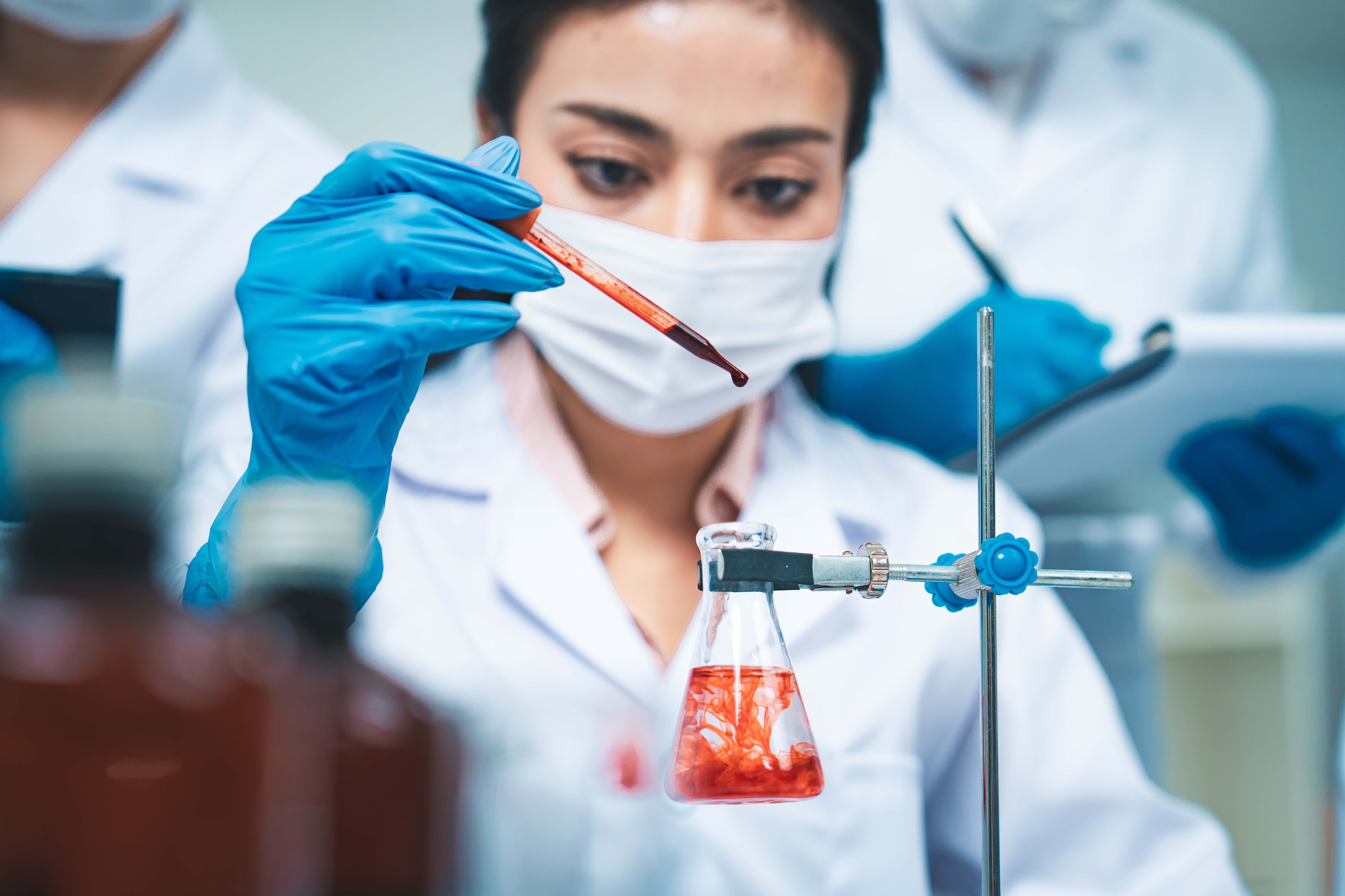 A female scientist with a mask pipetting a red solution into a lab flask.