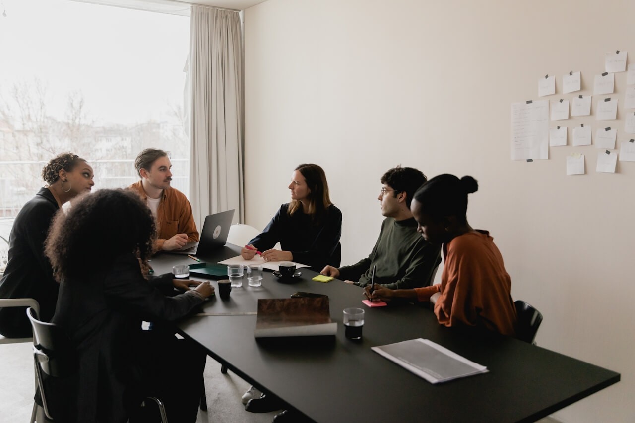 A diverse group of six people are sitting around a table in a well-lit conference room. They are engaged in a discussion, with laptops and papers on the table.