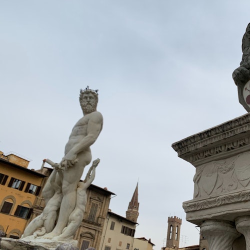 Une statue représentant une figure mythologique tenant un trident, située sur une place publique entourée de bâtiments historiques et d’un clocher.