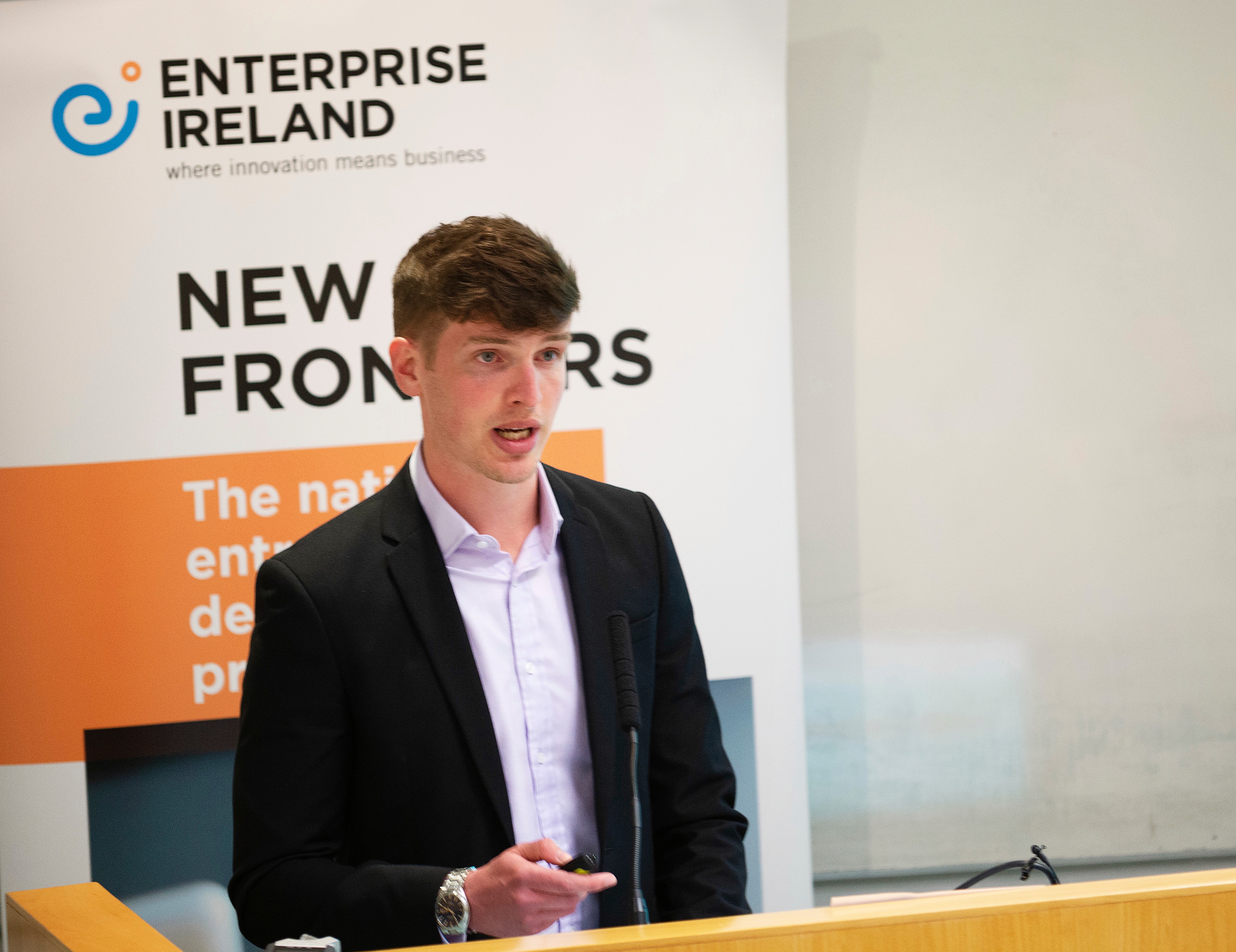 Ferdia Kenny presenting at the Enterprise Ireland New Frontiers Phase 3 final startup pitch day. He is wearing a black suit and is pitching in front of an Enterprise Ireland banner.
