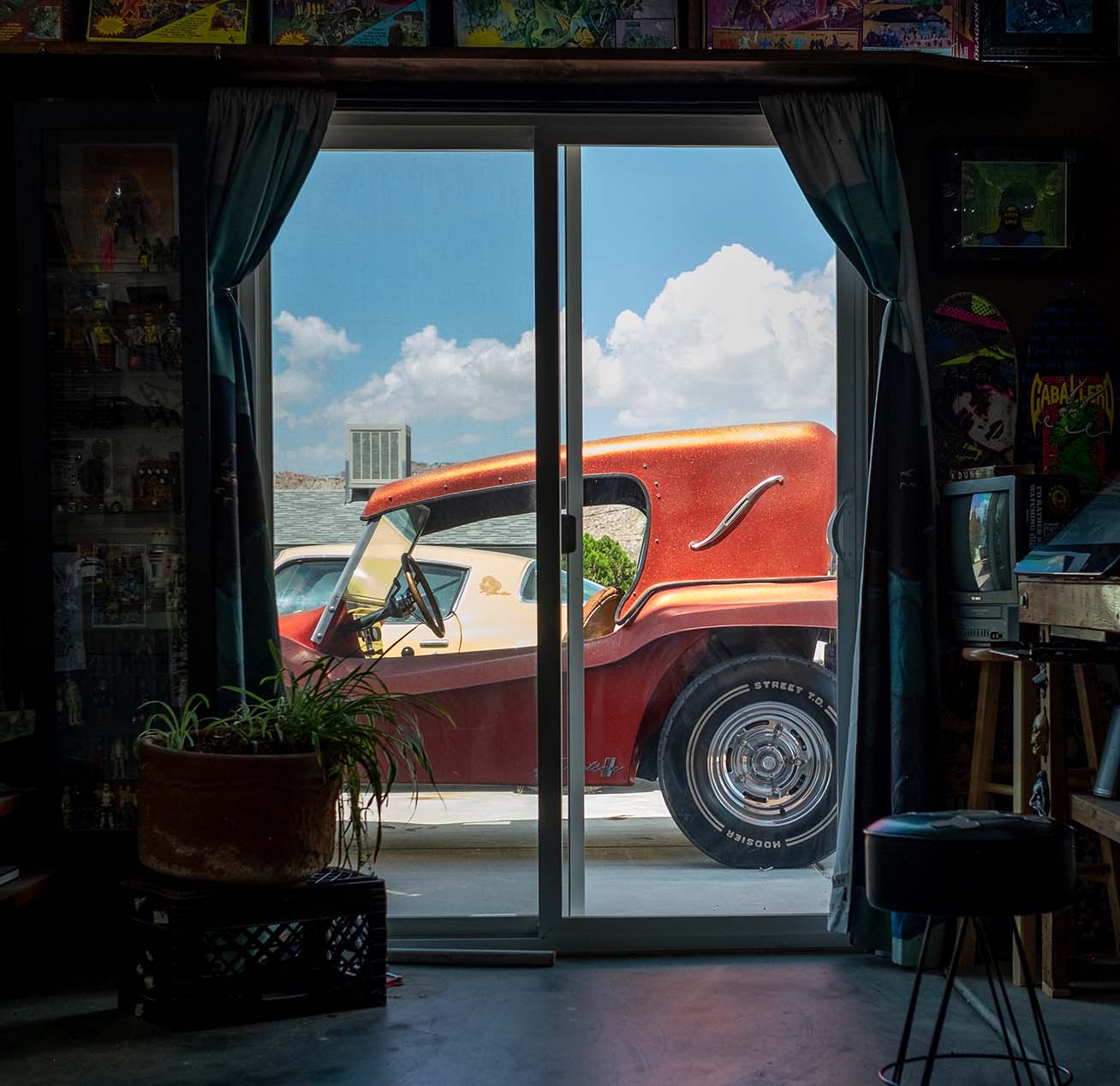 View through a sliding glass door showing a metallic orange Citation 2 buggy parked outside under a bright blue sky, framed by curtains and surrounded by art and collectibles inside the studio.