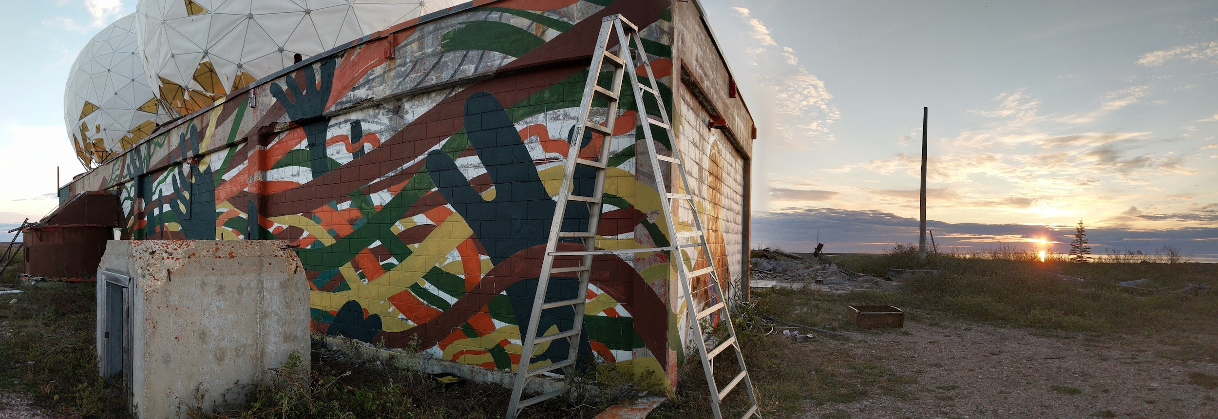 The sun sets low over the tundra beside the painted building, a ladder still leaning on its wall.