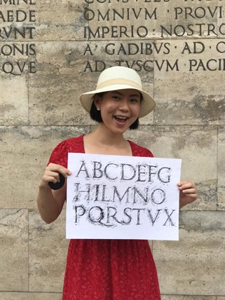 A photo of a girl holding a piece of paper with letters copied from a stone carved wall behind her.