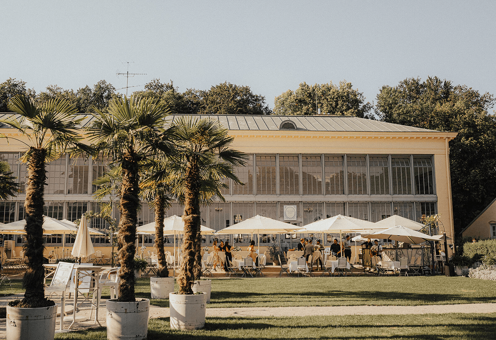 Outdoor restaurant terrace overlooking a tranquil lake at sunset. White tables and chairs are set with glassware. Sheer curtains add elegance. Calm ambiance.