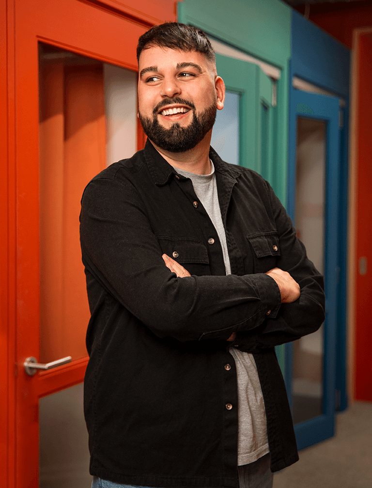Photo of Cameron Smith wearing and black open shirt shirt who is smiling and looking off camera with his arms folded.