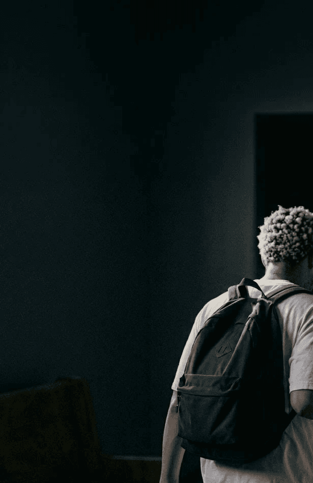 A person with curly hair walks down a dimly lit hallway wearing a casual t-shirt and a backpack, creating a mysterious and introspective atmosphere.