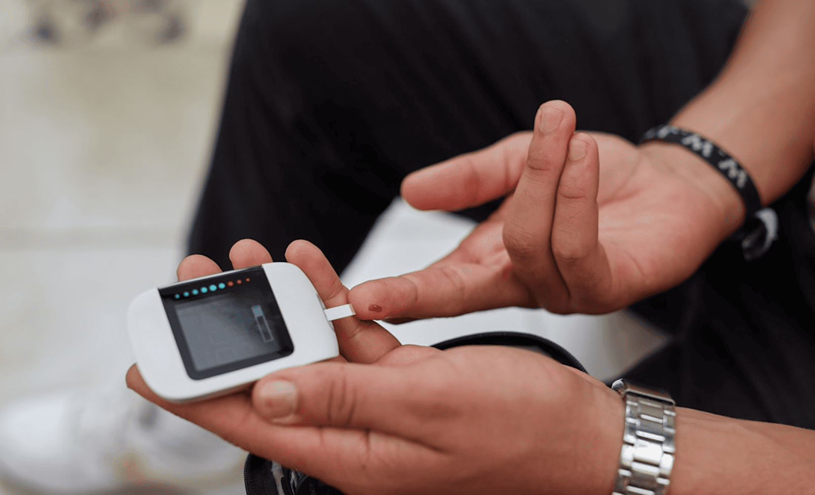Close-up of hands using a white digital blood glucose meter to check blood sugar levels, with a small drop of blood on the fingertip and a test strip inserted.