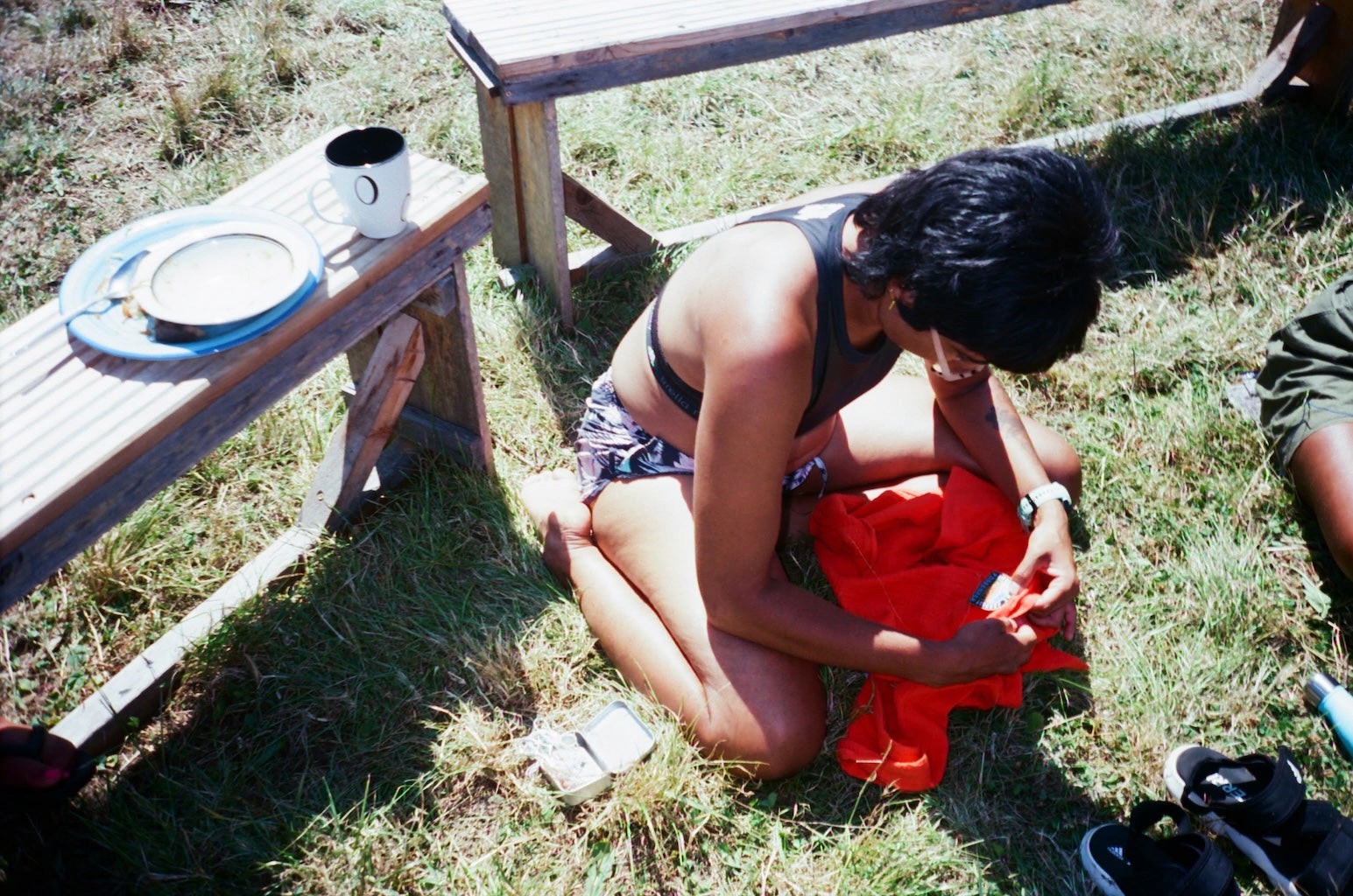 A person sits on the grass outside stitching a patch onto a top