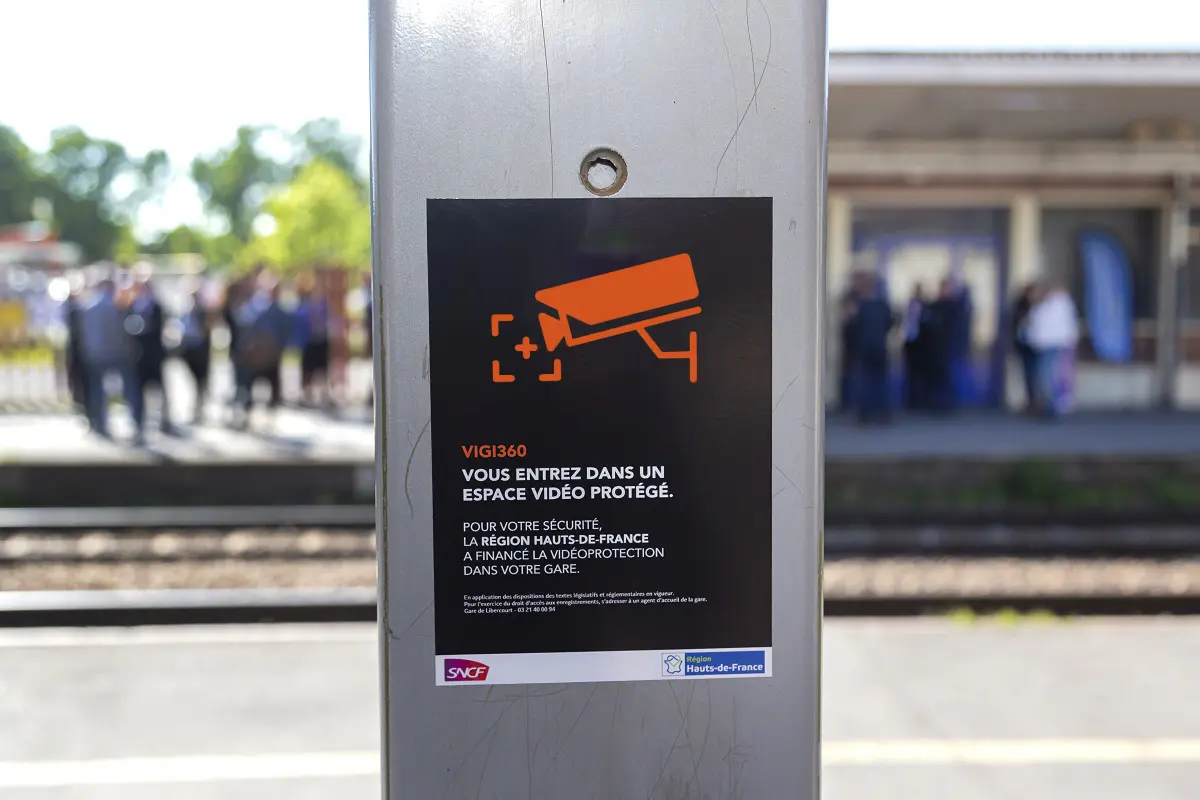 Black SNCF safety poster attached to a metal pillar on a train platform, featuring the orange camera pictogram and text about entering a protected video-monitored space.