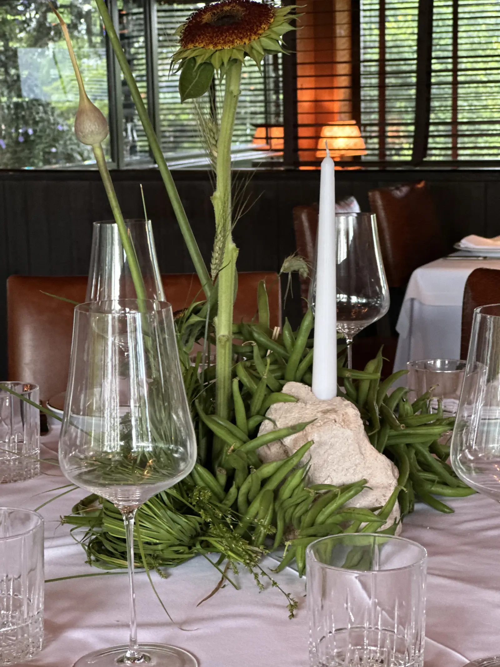 Arreglo orgánico de mesa con abundantes chauchas verdes frescas enmarcando una vela blanca cilíndrica.