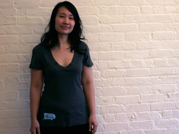 A person with long dark hair stands against a white brick wall, wearing a dark gray v-neck t-shirt and dark pants. The t-shirt has a small, light blue rectangular tag near the bottom left. The person’s expression is neutral, and they appear to be looking directly at the camera.