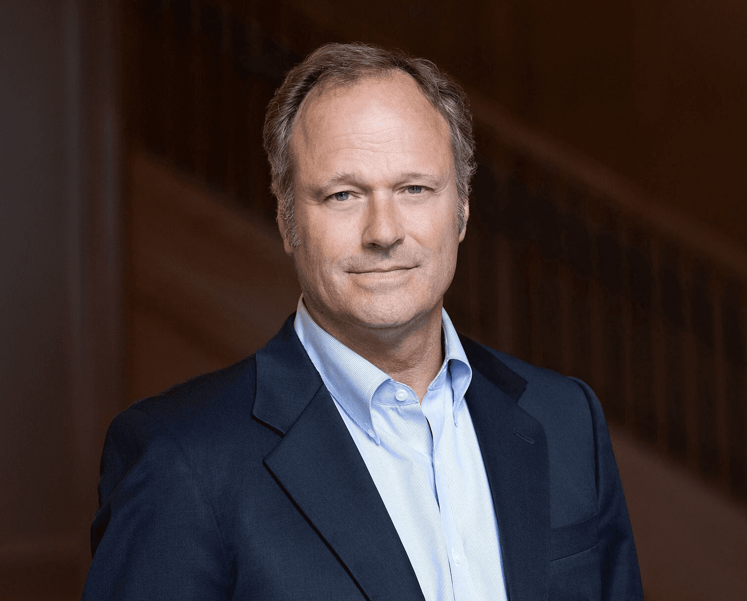 A professional headshot of a man in a suit, smiling against a blue background.