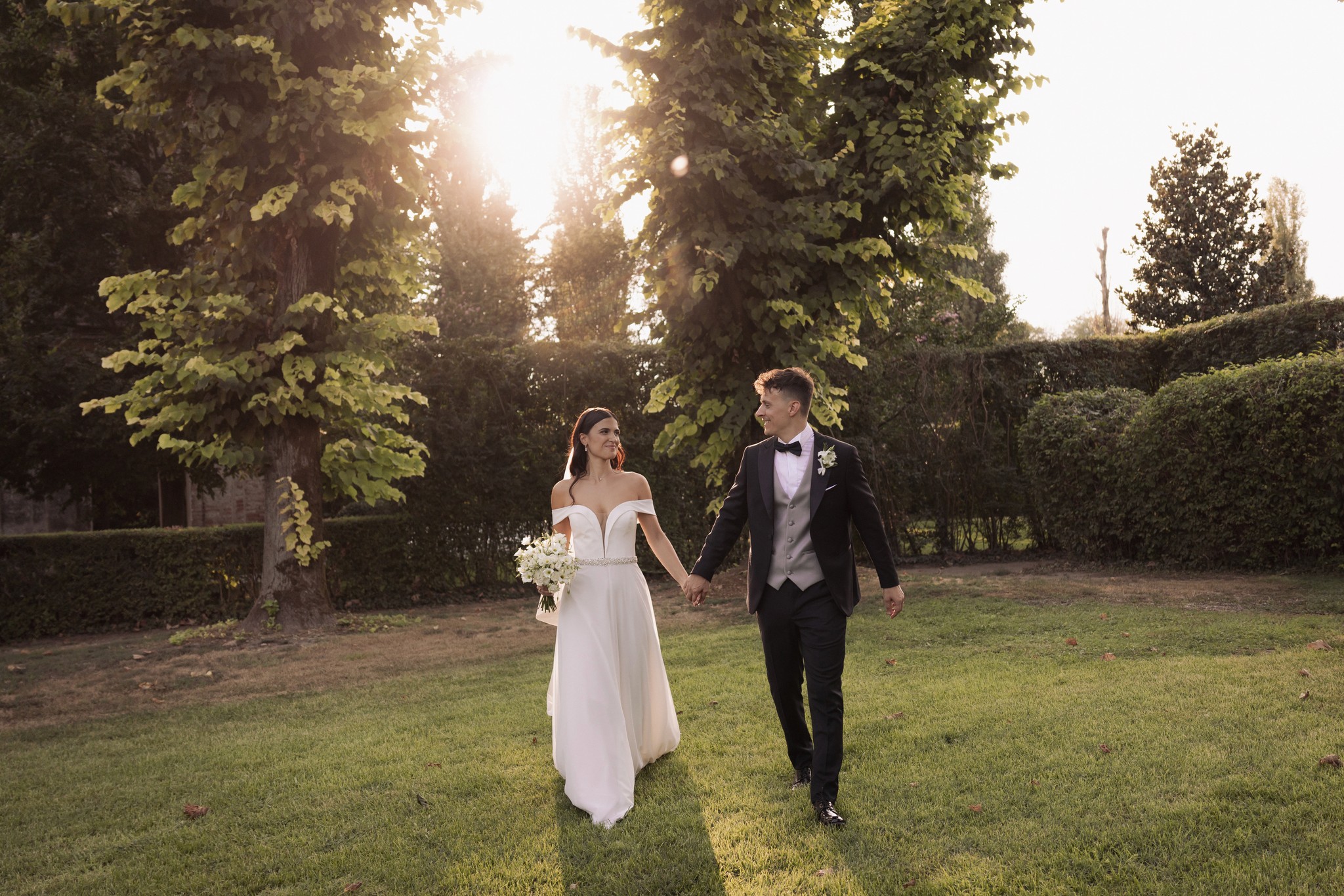 Sposi camminano mano nella mano nel giardino di Villa Negri al tramonto, lei in bianco, lui in smoking tra alberi