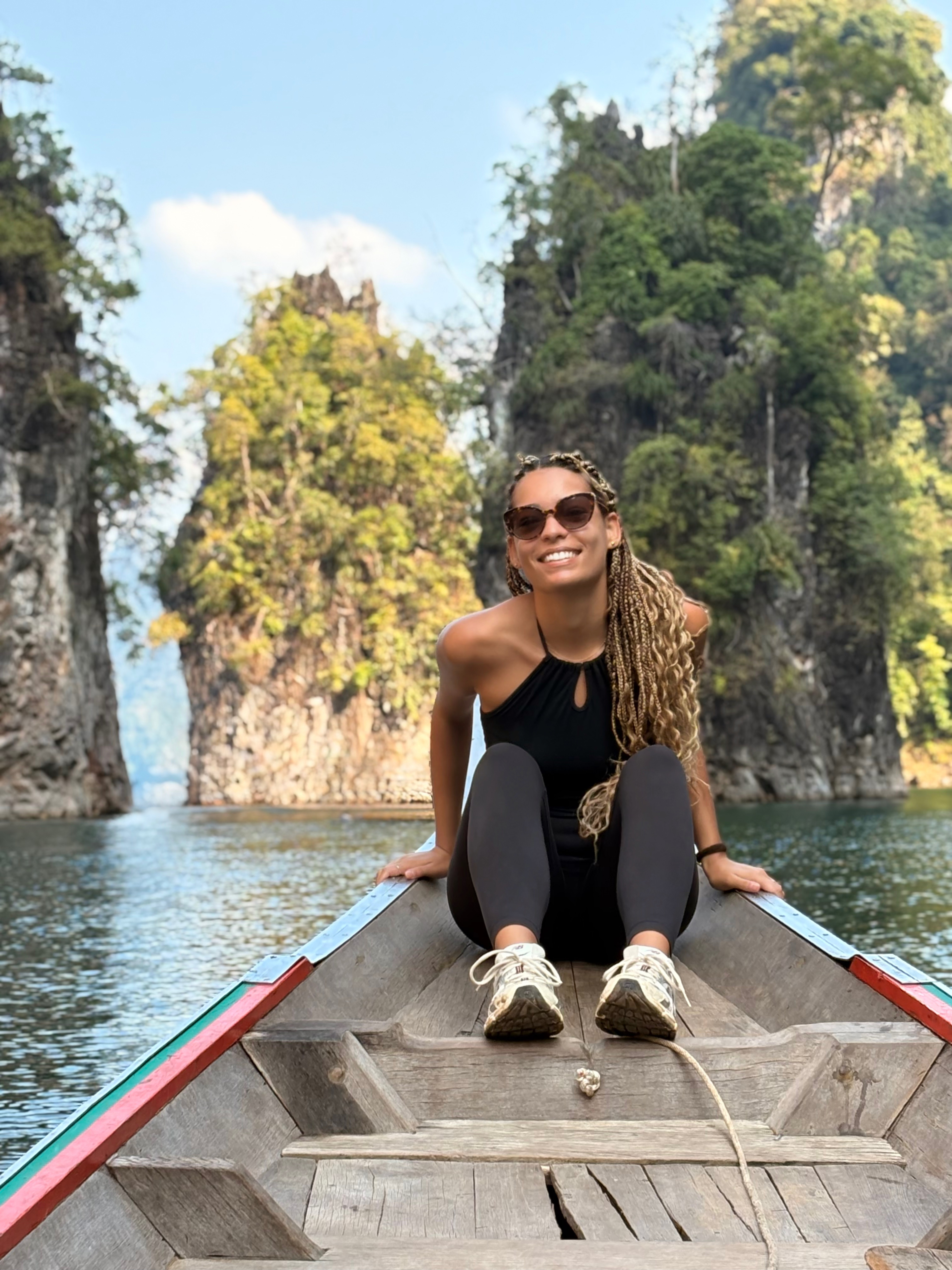 A smiling woman sits at the end of a narrow wooden boat in a tropical sea surrounded by karsts