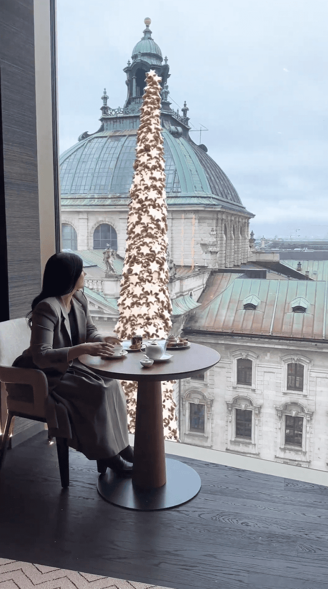 A woman has tea by a window with a view of a massive star-covered Christmas tree and a historic dome
