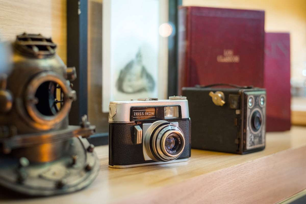 A vintage silver and black camera sits on a wooden surface next to an old diving helmet and two red books.