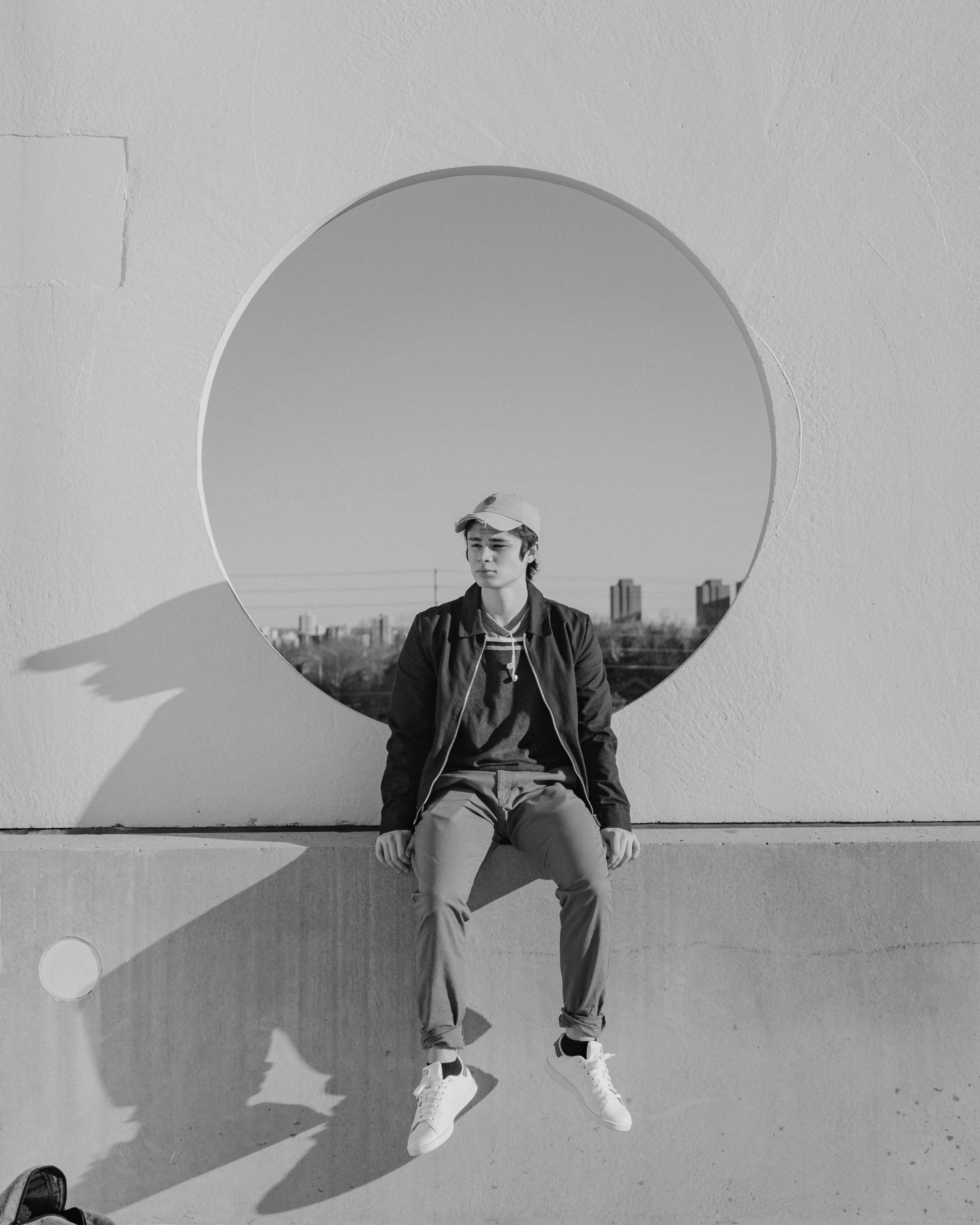 A person in casual attire sits against a wall with a circular shadow, appearing relaxed and playful.