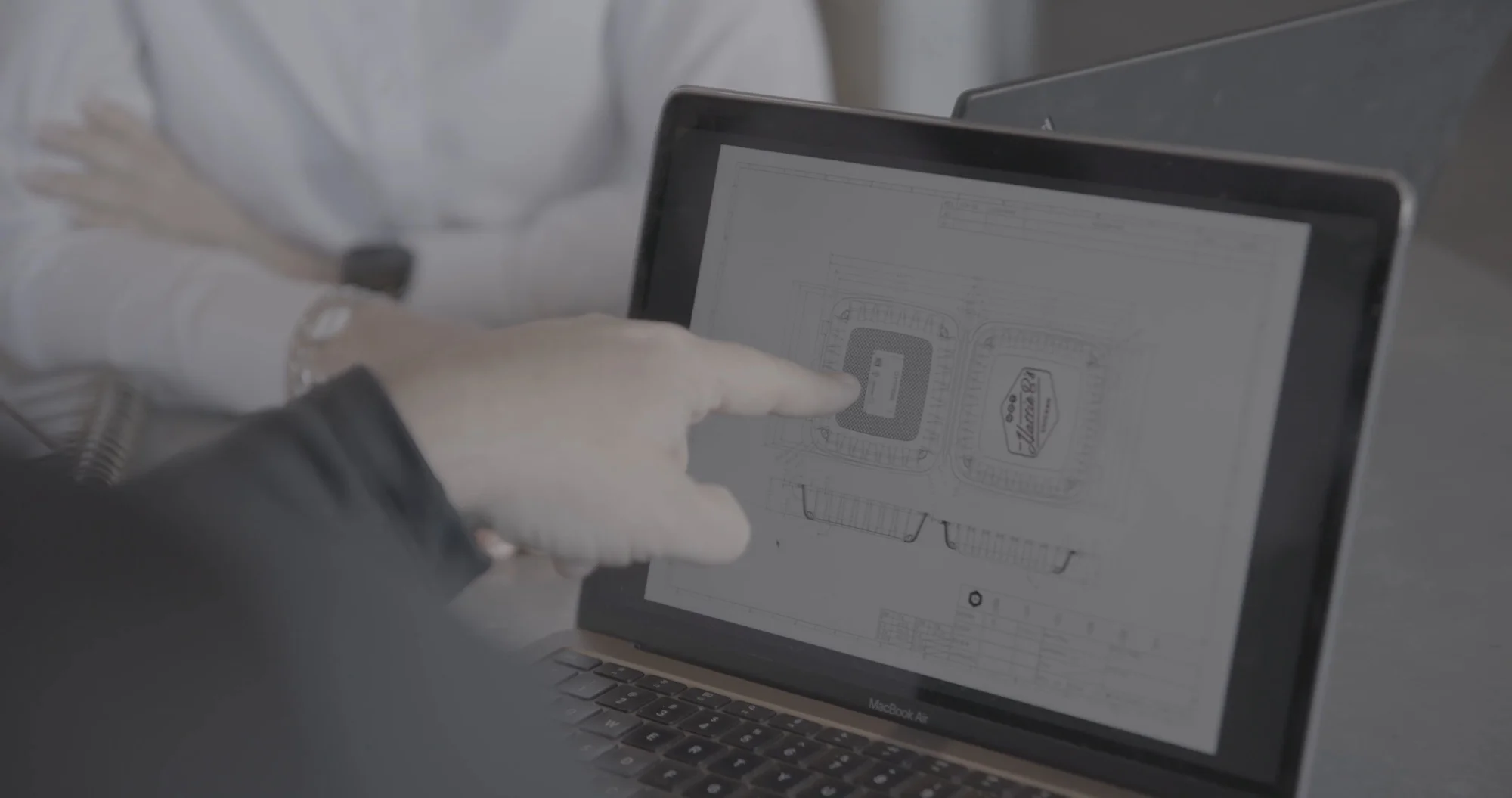 A person wearing gloves points at a blank screen of a tablet or device. The background is softly blurred.