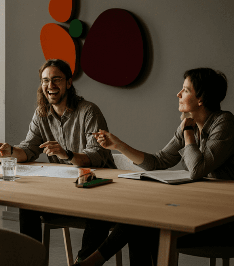 A group of three colleagues engaged in a lively discussion around a wooden conference table in a modern, well-lit office space, with papers, notebooks, and a glass of water placed on the table.