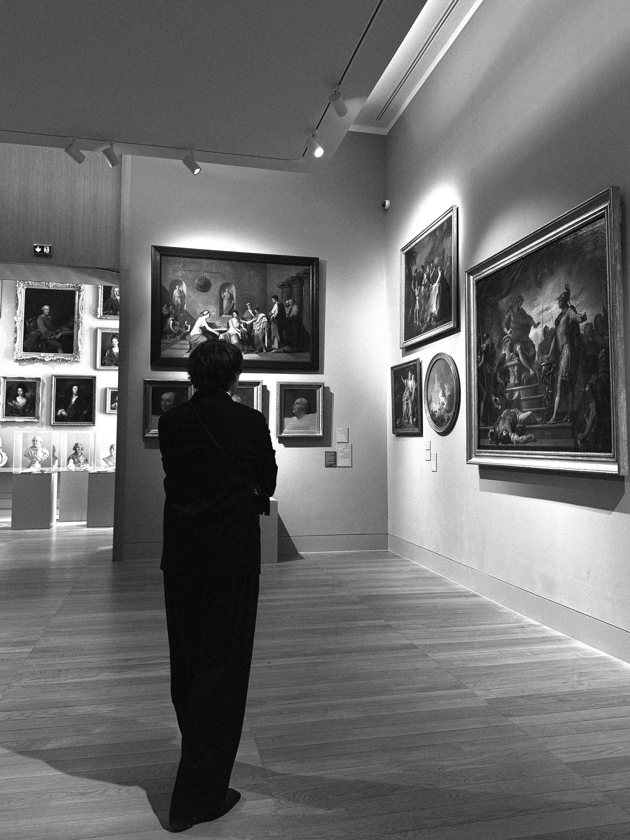 Maximilien, fondateur du studio, se tient dans une vaste galerie de musée d'art, contemplant un mur orné de diverses peintures classiques dans des cadres ornés, sous un éclairage doux et focalisé.