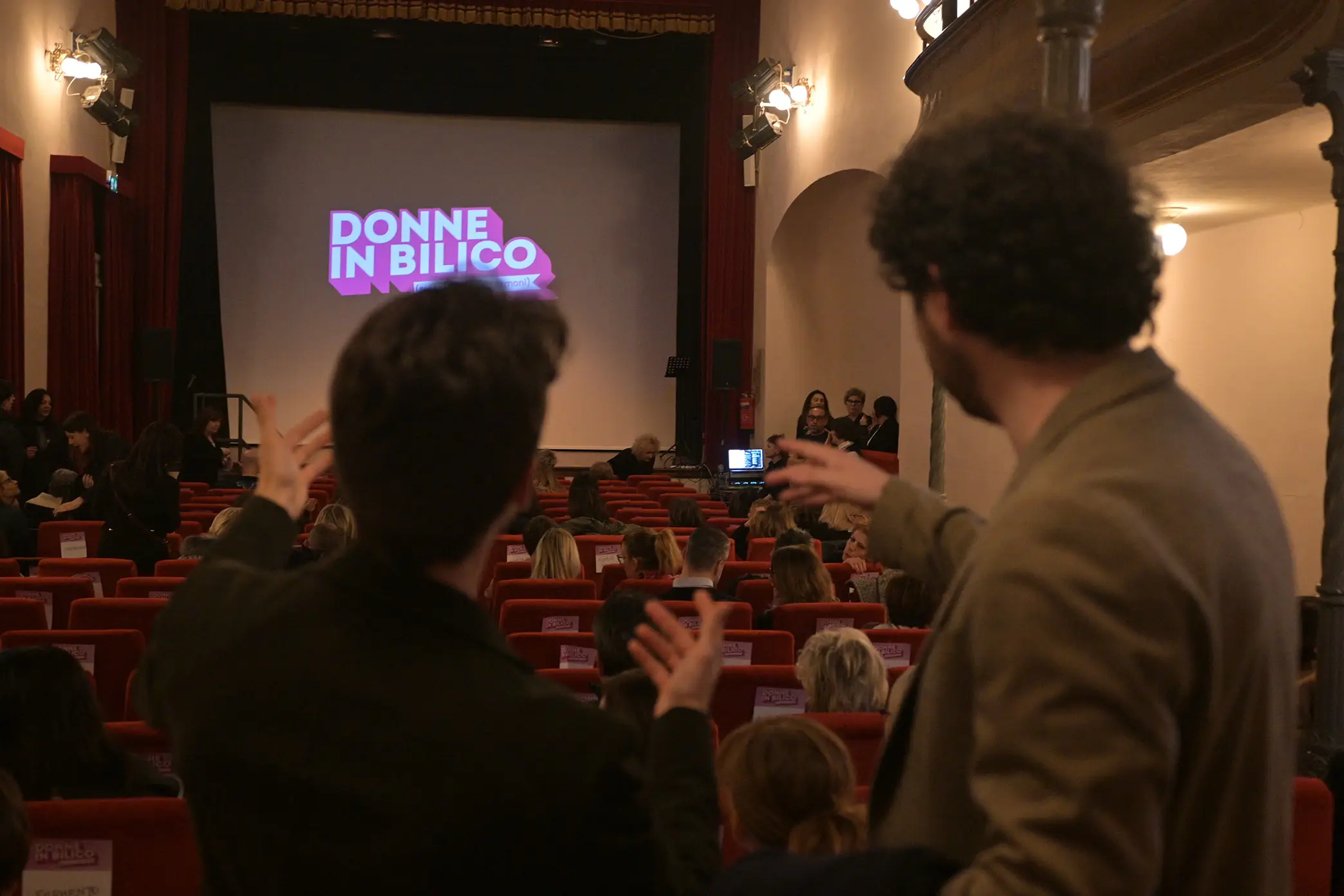 Due ragazzi di spalle nella sala del teatro verdi di Forlimpopoli che indicano lo schermo con il logo Donne in Bilico proiettato