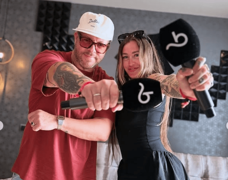 a photo of a man in a red tshirt anda hat and a woman in all black, sunglasses on her head and long brown hair (anna) holding booksy microphones to the camera posing for the photo