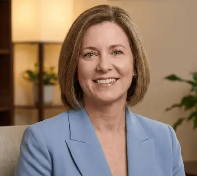 A smiling woman with shoulder-length hair, wearing a light blue blazer, is sitting in a well-lit room.