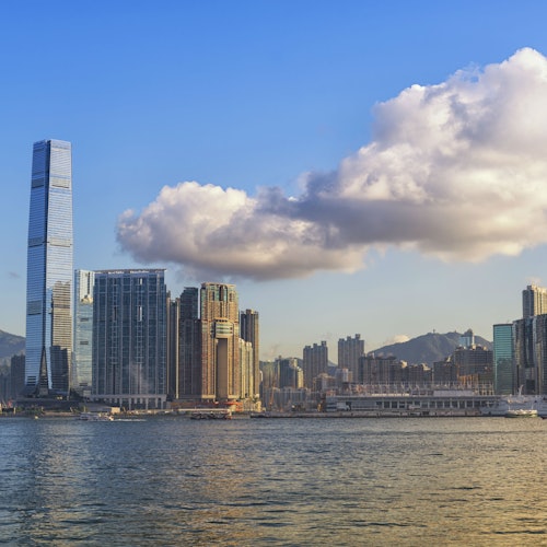 Magas felhőkarcolók egy vízpart mellett, háttérben hegyekkel és egy nagy, bolyhos felhővel a kék égbolt alatt.