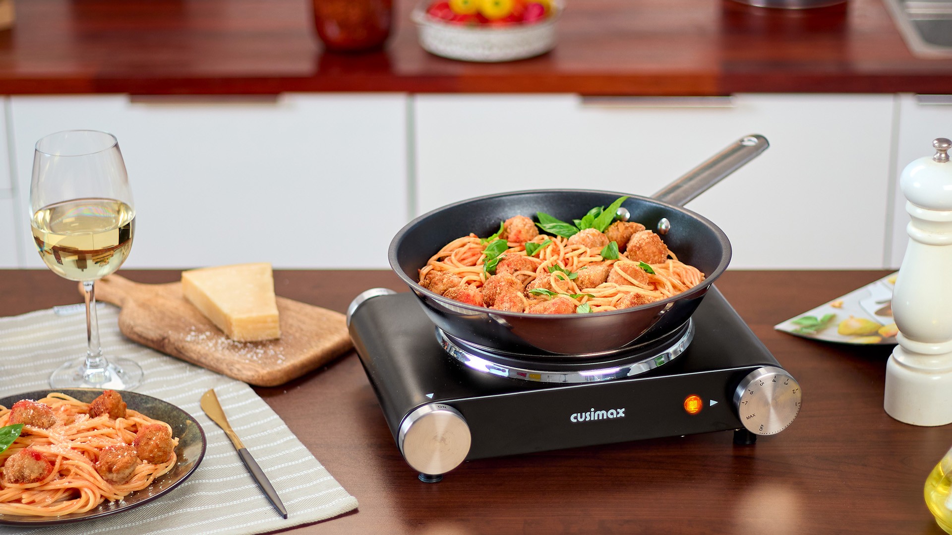 A plate and skillet filled with spaghetti and meatballs, garnished with fresh basil, sit on a kitchen counter alongside a glass of white wine, a block of Parmesan cheese, and a pepper mill.