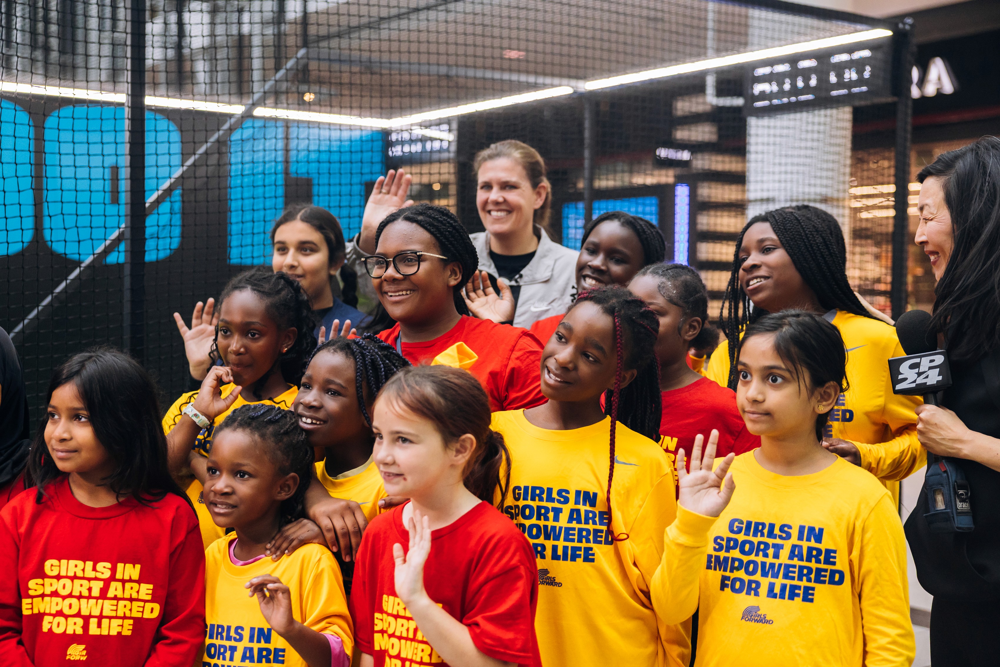 A joyful group of girls, diverse in appearance, wear red and yellow shirts with "Girls in Sport Are Empowered for Life," smiling and raising hands.