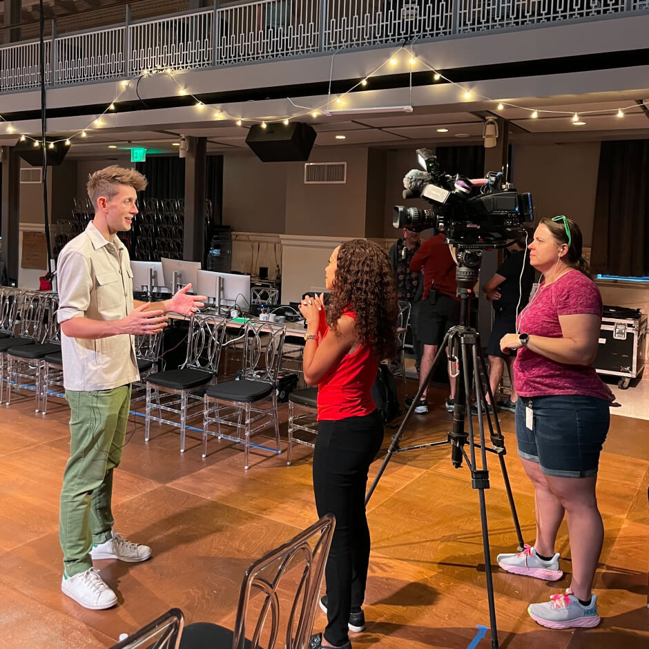 A man is being interviewed by a woman with a microphone while a camerawoman films them inside a large hall with string lights and empty chairs.