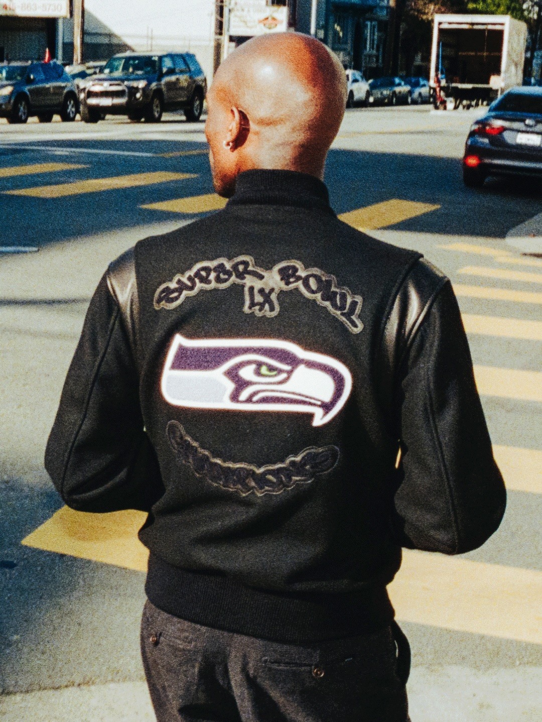 A shot of the back of the Fanatics X Apexer Seahawks Varsity Jacket on a model walking across the street