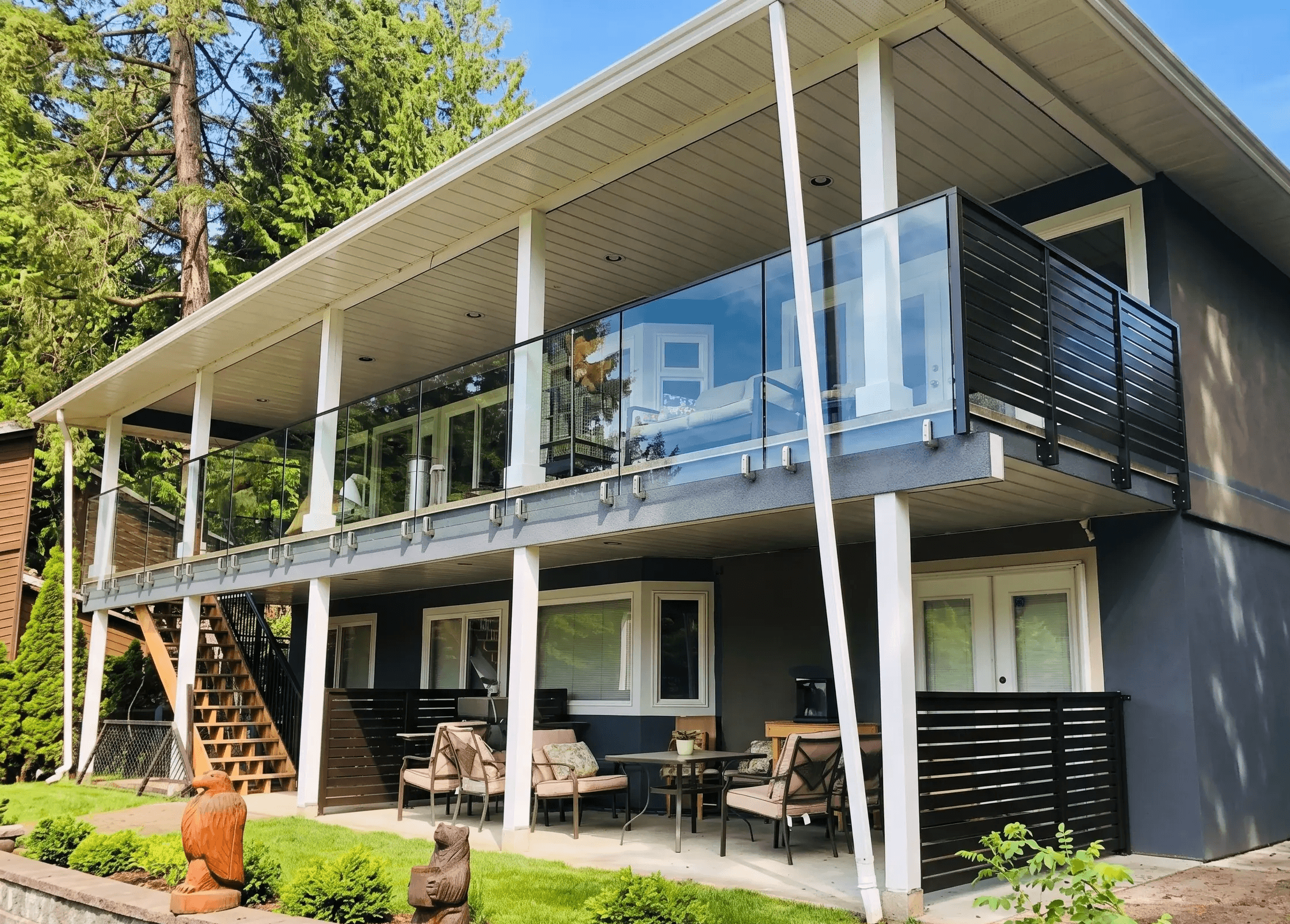 Exterior shot of a two-story home featuring a spacious upper-level balcony enclosed by clear glass railings along the front and dark horizontal privacy slats on the side. The deck is supported by white pillars over a ground-level covered patio furnished with outdoor seating. In the foreground, there is a low block retaining wall with manicured green shrubs and two carved wooden statues of an eagle and a bear.