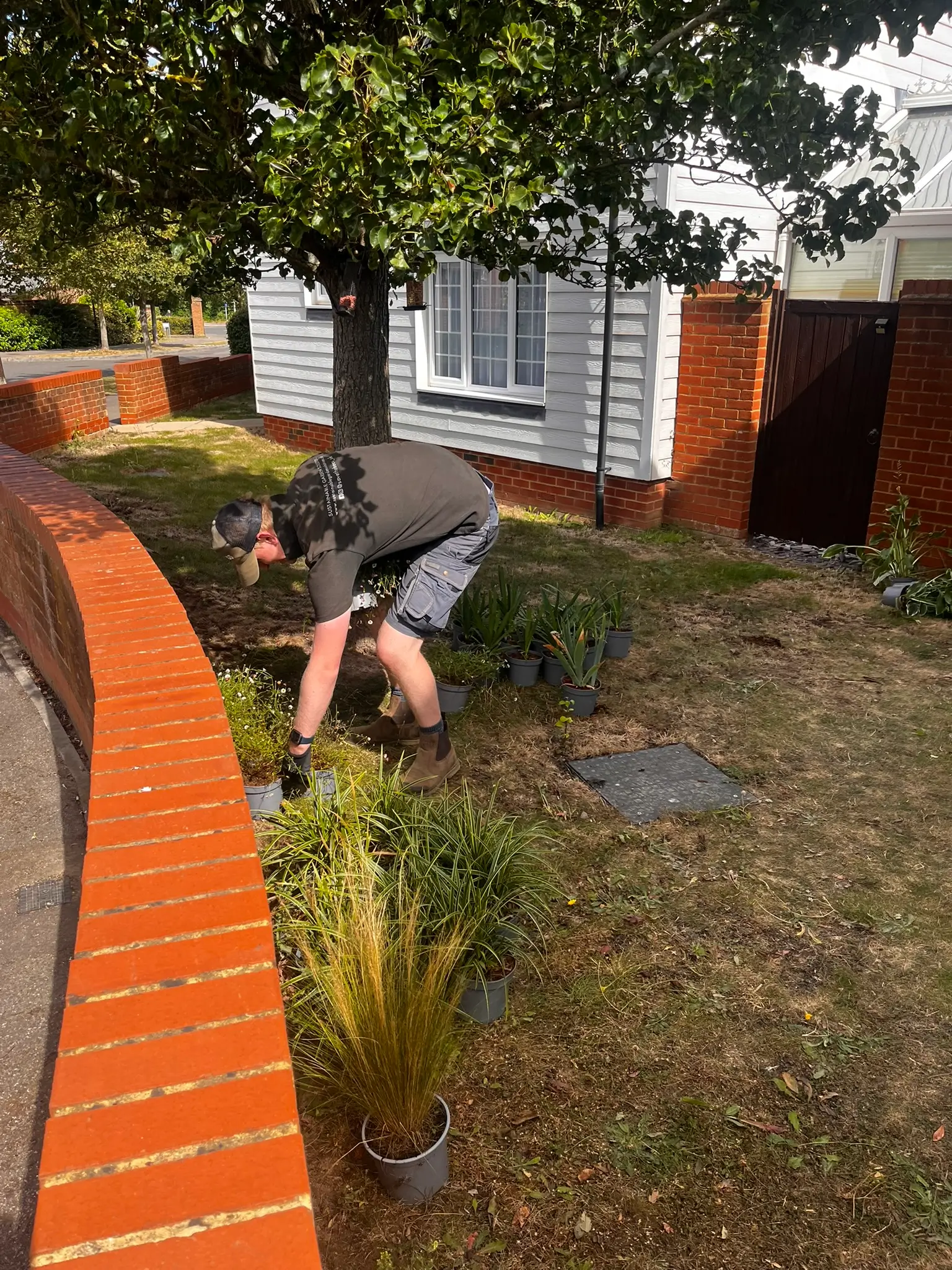 A person is bending down to tend to plants in a garden next to a brick wall and a house in the background.