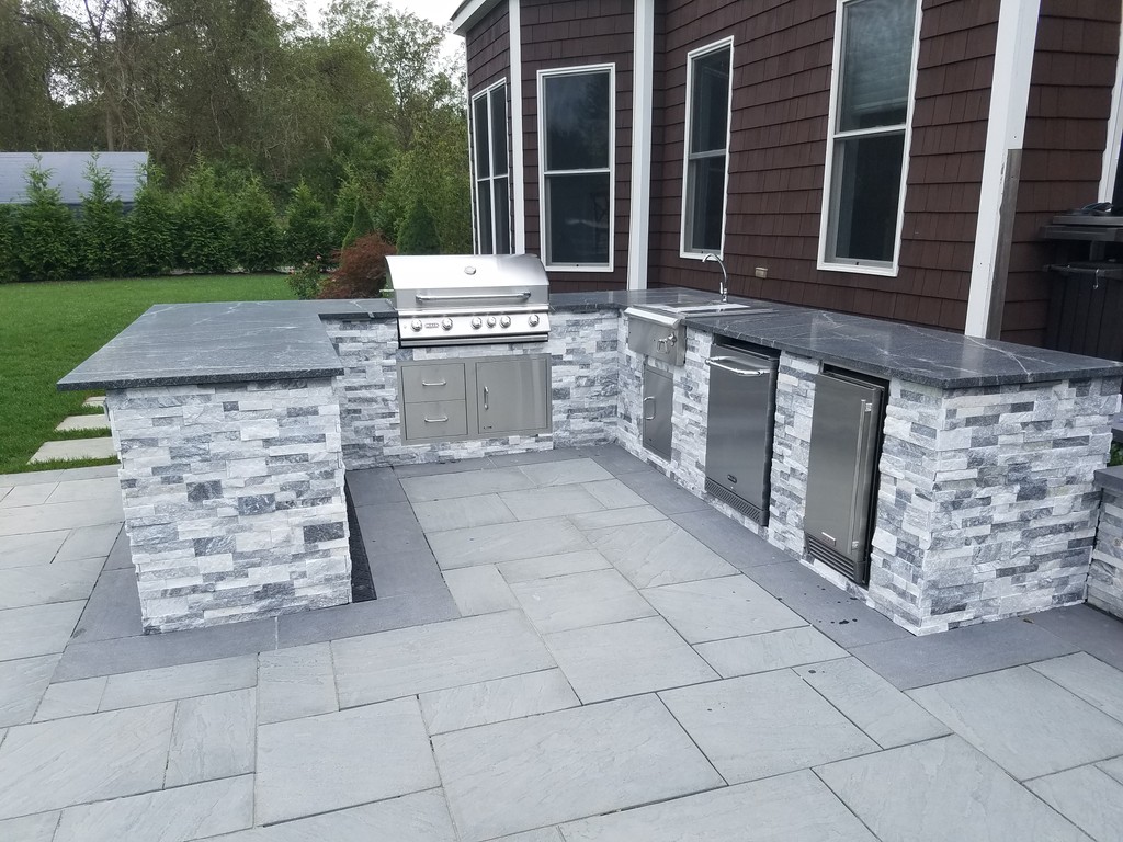 Modern gray-stone outdoor kitchen with built-in grill, sink, and fridge
